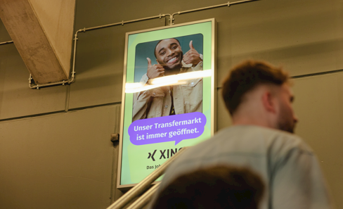 A digital OOH display showing a young black man grinning with two thumbs up and the text in a speech bubble: “Unser Transfermarkt ist immer geöffnet.”